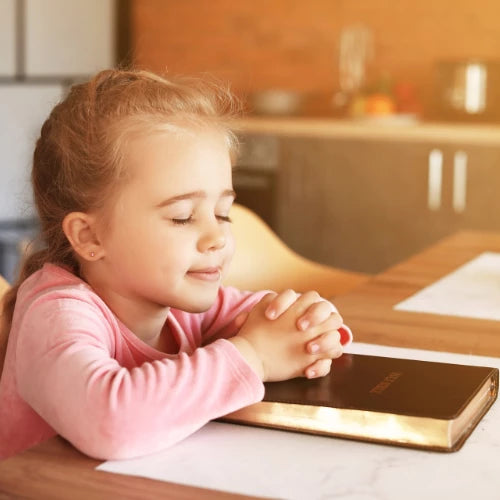 Little Girl Praying with Holy Bible at Table with Eyes Closed and Hands Folded